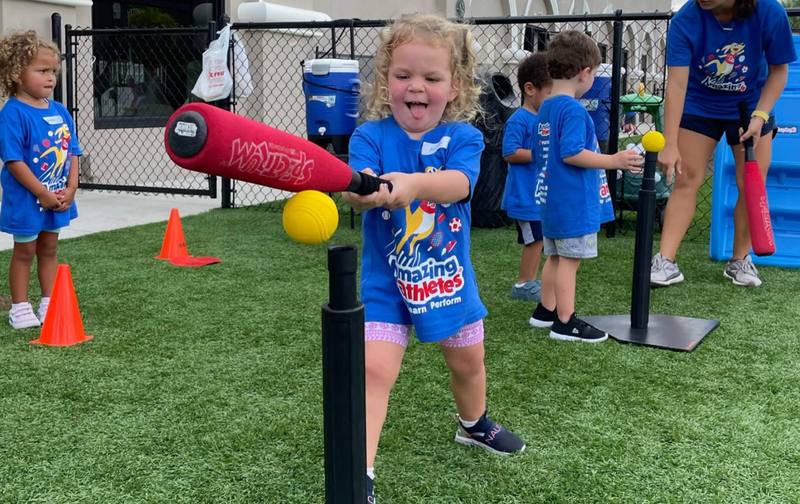 A preschool girl in an Amazing Athletes shirt swinging a foam bat at a ball on a tee
