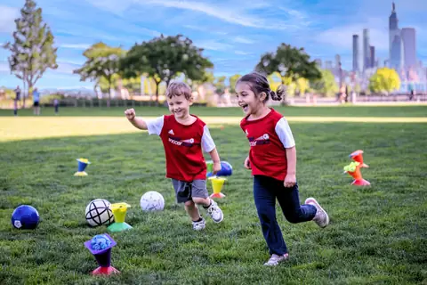 Two preschoolers in Soccer Stars jerseys racing across a park lawn with the city skyline behind them