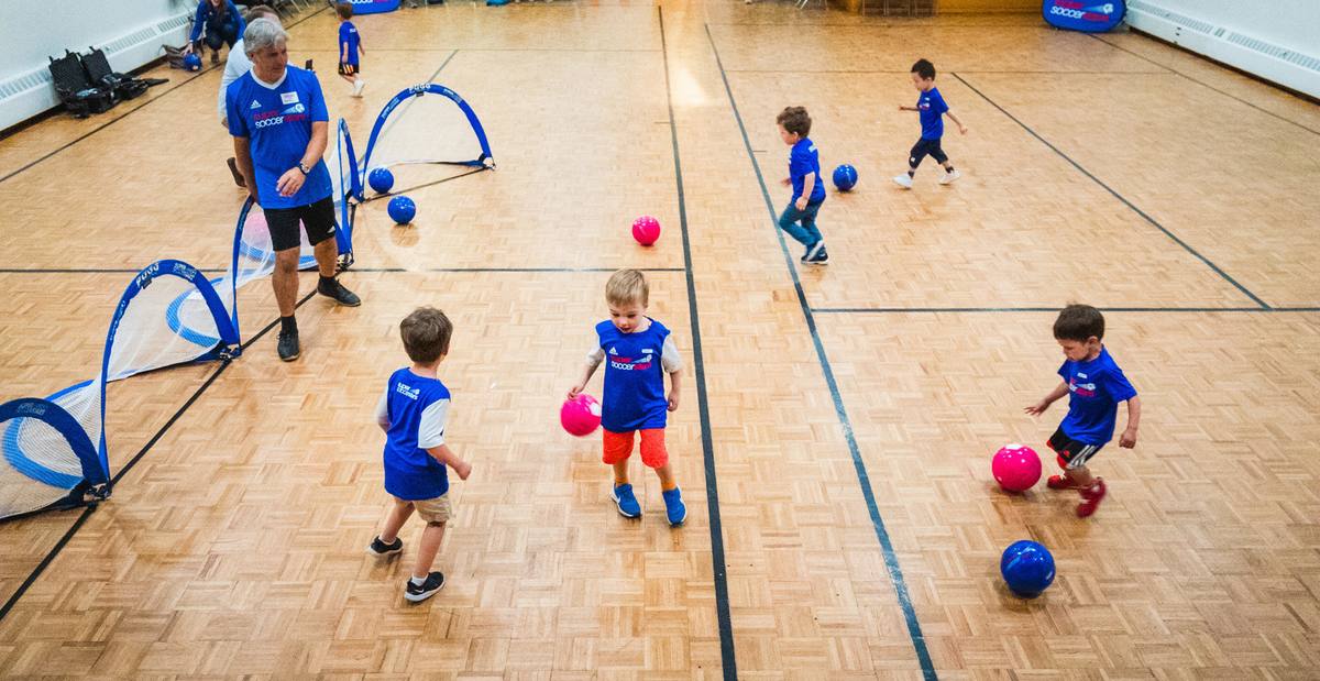 A Soccer Stars coach leading five young children dribbling soccer balls around pop-up goals in a school gym