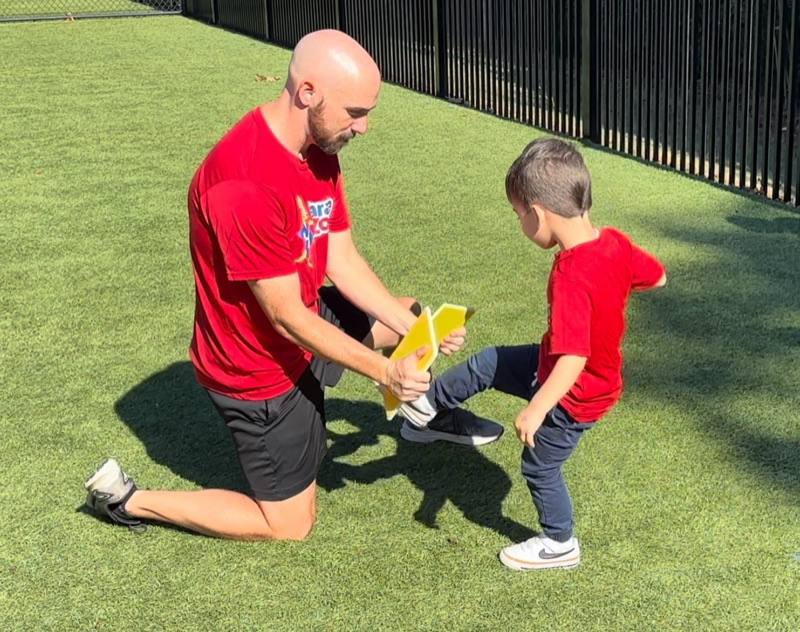 A Karate Zoo instructor kneeling and holding a kick pad while a young boy practices a kick