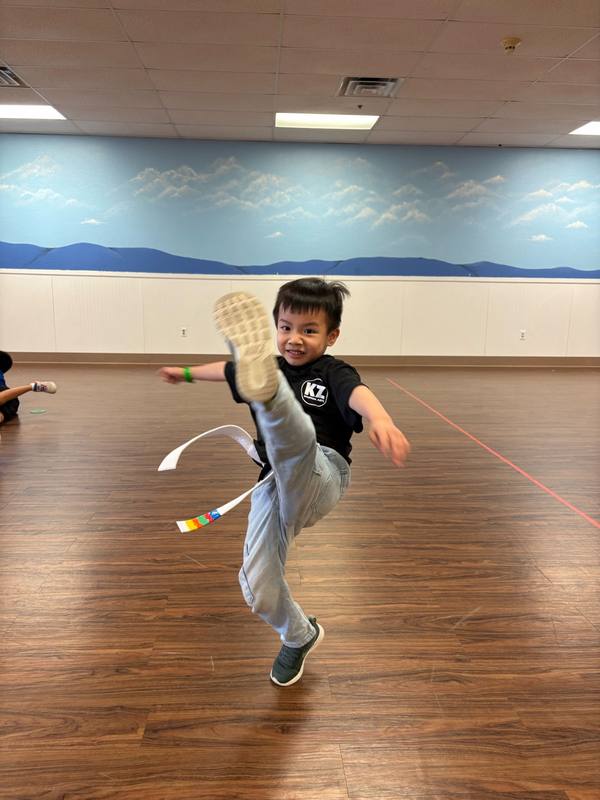 A boy in a Karate Zoo shirt in the middle of a high kick in a multipurpose room