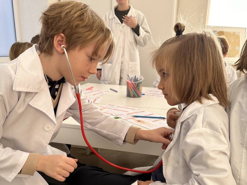 Two children in white lab coats at a classroom table, one listening to the other's heartbeat with a stethoscope