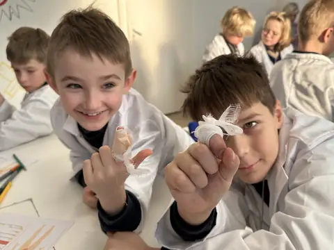 Two grinning boys in lab coats holding up fingers they bandaged during a Little Medical School lesson