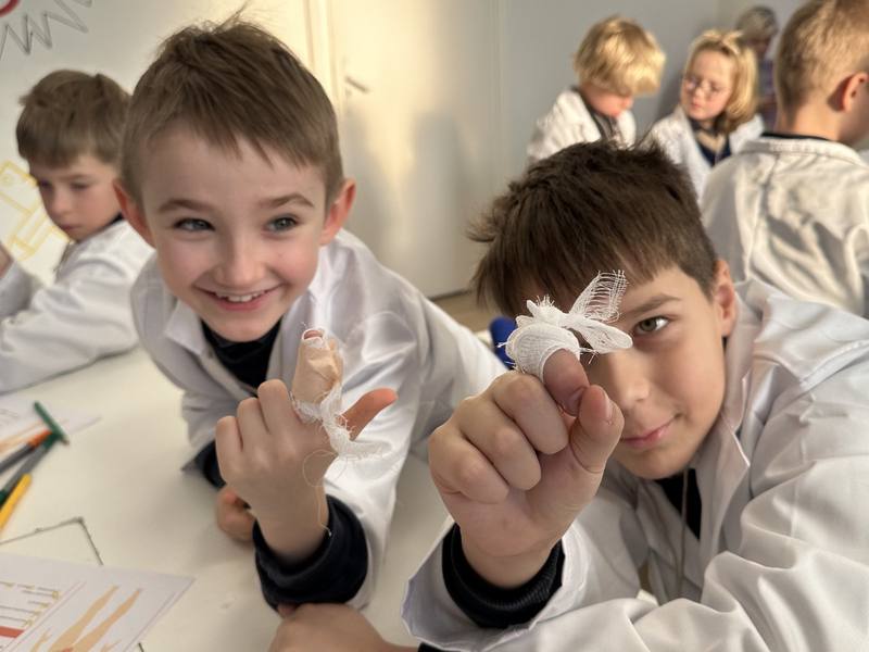 Two grinning boys in lab coats holding up fingers they bandaged during a Little Medical School first aid lesson