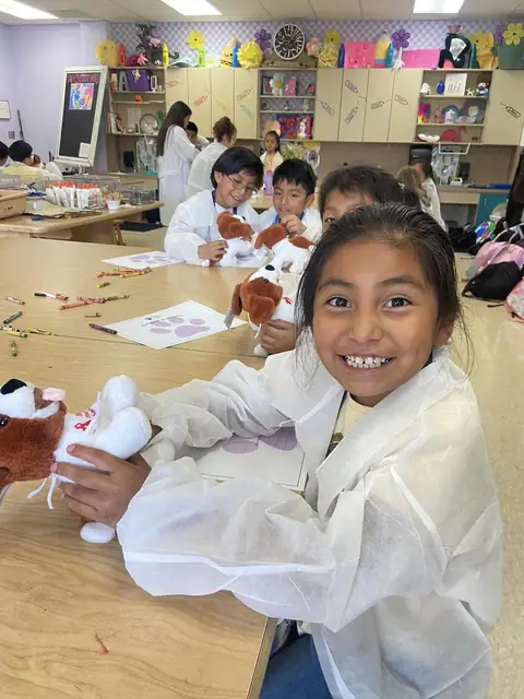 A smiling girl in a lab coat examining a plush dog at a classroom table