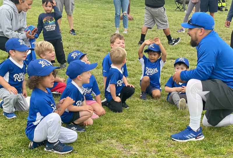 A Little Rookies Baseball coach kneeling on the grass talking with a circle of small children in caps and jerseys