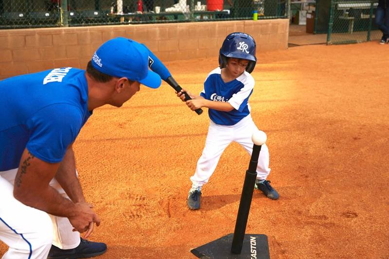A Little Rookies Baseball coach crouching beside a batting tee as a small boy in a helmet takes a swing