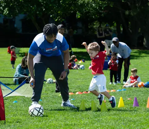 A Soccer Stars coach rolling a ball while a toddler jumps over cones in a park