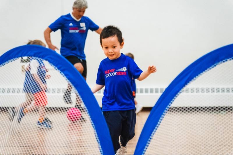 A smiling preschooler in a Soccer Stars jersey running between two pop-up goals in a gym while a coach follows behind