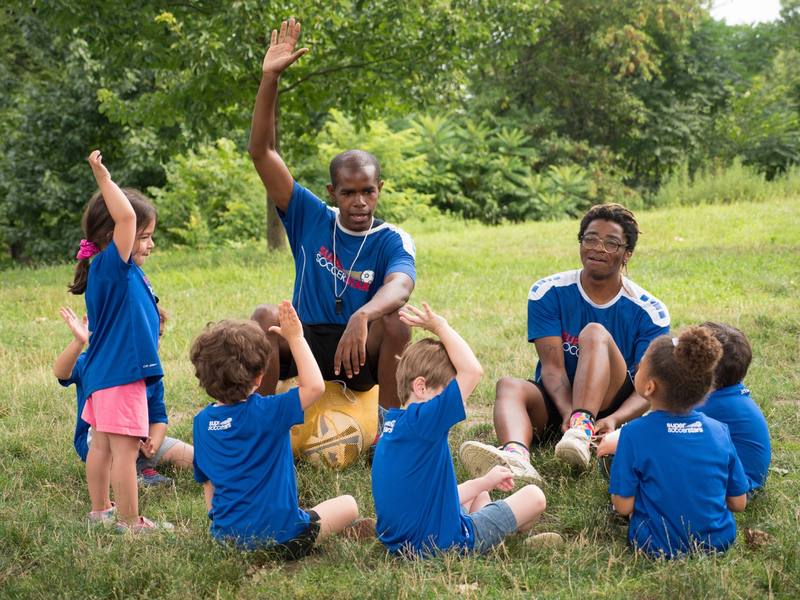 Two Soccer Stars coaches sitting in the grass with a circle of young children, everyone raising a hand